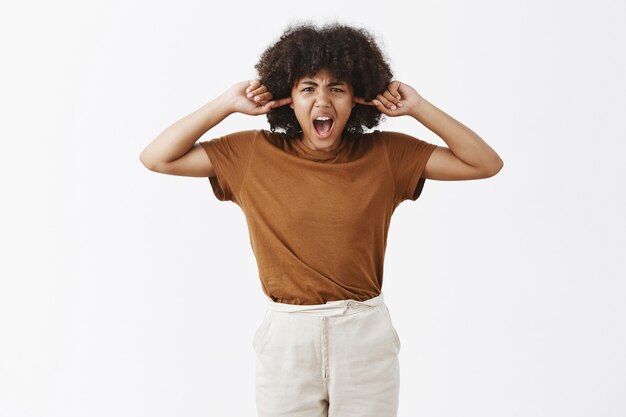 Portrait of bothered and annoyed displeased unhappy African American woman with afro hairstyle screaming while closing ears with index fingers during argument