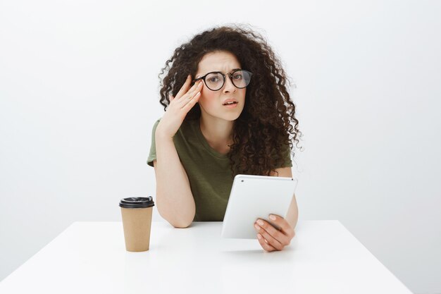 Portrait of bothered clueless curly-haired european woman in black glasses, sitting at table, holding smartphone and drinking coffee or tea, holding hands on temples