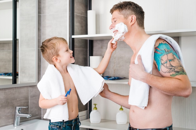 Foto gratuita ritratto di un ragazzo che applica la schiuma da barba sul viso di suo padre in bagno