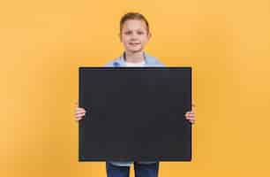 Free photo portrait of a boy holding black chalkboard standing against yellow background