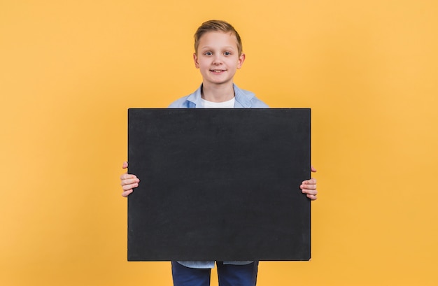 Portrait of a boy holding black chalkboard standing against yellow background