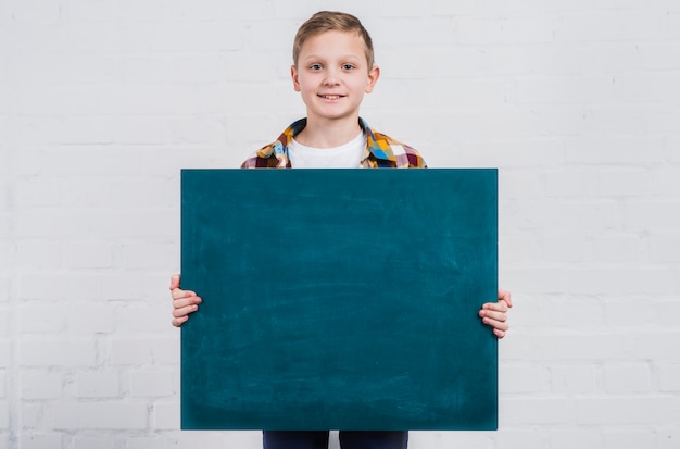 Portrait of a boy holding blank chalkboard standing against white brick wall