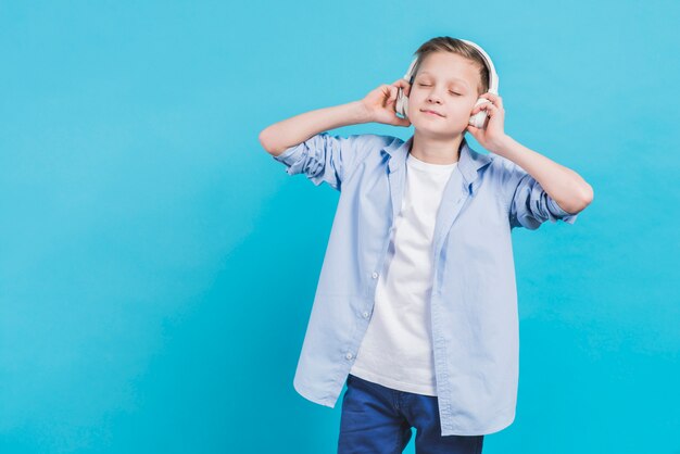 Portrait of a boy listening music on white headphone against blue background