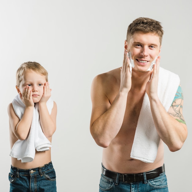 Portrait of a boy touching his cheeks standing near his smiling father applying shaving foam on face