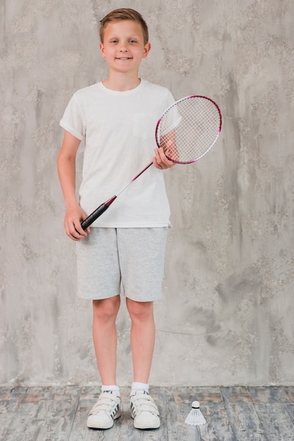 Portrait of a boy with racket and shuttlecock standing in front of concrete wall