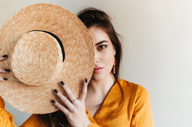 Portrait of brown-eyed girl covering face with straw hat. Beautiful lady in bright outfit