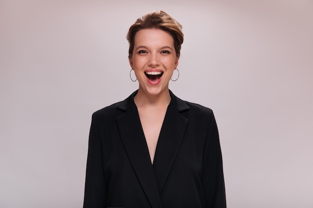 Portrait of brown eyed lady in dark jacket on isolated background. Attractive short-haired woman in black suit laughs and looks into camera on white backdrop