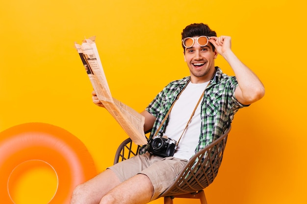 Портрет кареглазого мужчины, снимающего очки и читающего газету, сидя на стуле на оранжевом фоне