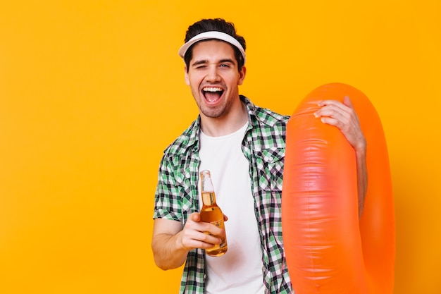 Portrait of brunet guy in white cap pointing to camera. Man with rubber ring holding beer.