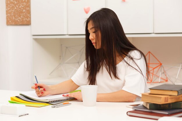 Free photo portrait of brunette asian student writing something in her exercise-book and drinking delicious cup of tea or coffee in library.