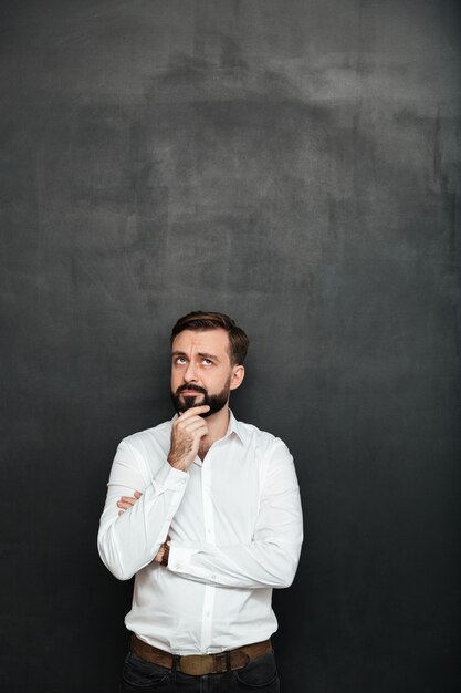 Portrait of brunette bearded man in white shirt touching his chin thinking or remembering over dark gray