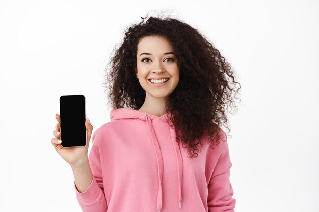 Портрет кудрявой брюнетки, показывающей приложение на телефоне, пустой экран смартфона, улыбающейся, рекомендующей скачать приложение, стоящей на белом фоне