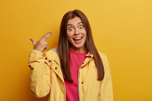 Portrait of brunette European female has surprised expression, laughs and makes finger gun pistol gesture, shoots in temple, foolishes around as being bored for long waiting, wears yellow jacket