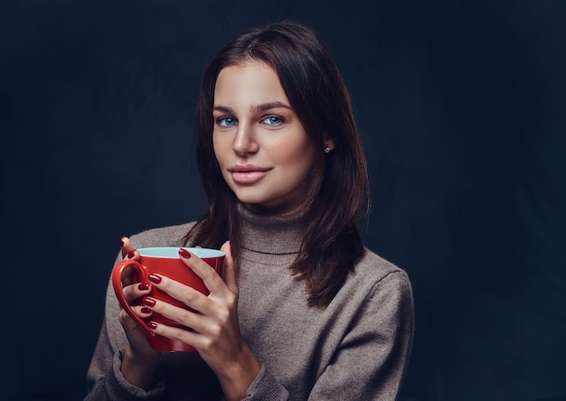 Portrait of brunette female dressed in a brown long neck jacket holds the red coffee cup.