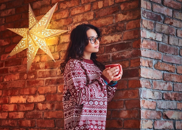 Portrait of brunette female dressed in a red sweater drinks coffee over the wall of a brick.