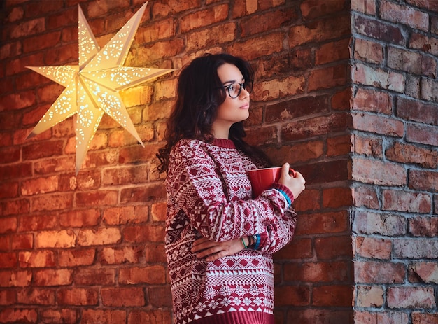 Portrait of brunette female dressed in a red sweater drinks coffee over the wall of a brick.