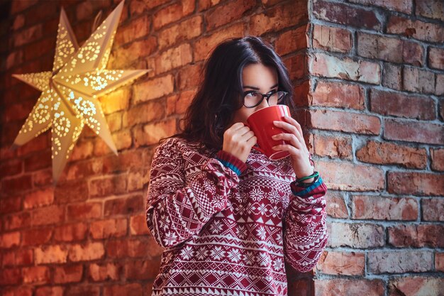 빨간 스웨터를 입은 갈색 머리 여성의 초상화는 벽돌 벽 너머로 커피를 마신다.