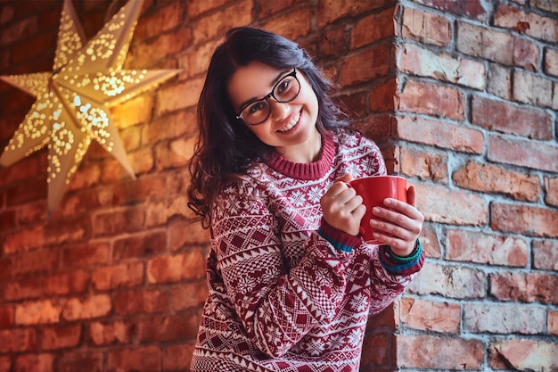 Portrait of brunette female dressed in a red sweater drinks coffee over the wall of a brick.