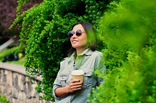 サングラスをかけたブルネットの女性の肖像画は、夏の公園で外出先でコーヒーを飲みます。