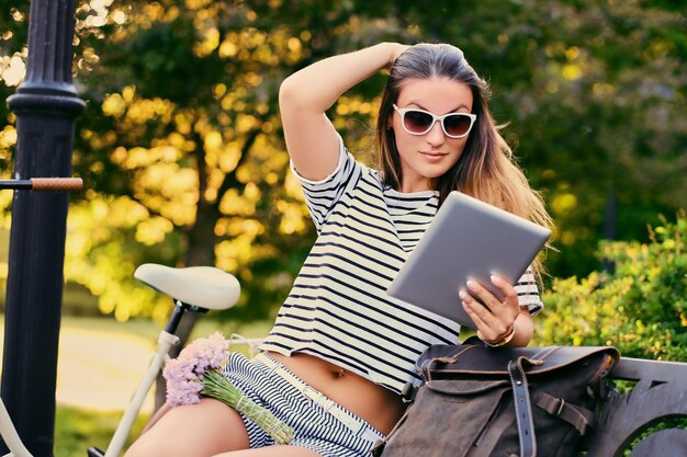 Portrait of brunette female using tablet PC with city bicycle in a park on background.