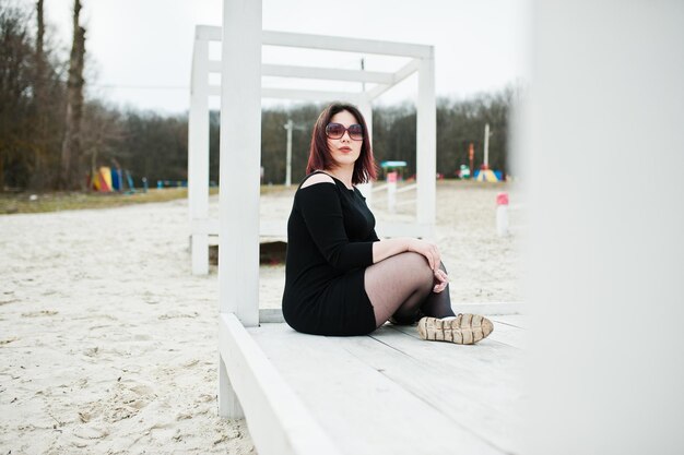 Portrait of brunette girl in black dress sitting at white wooden construction