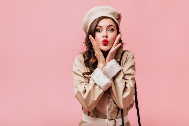 Portrait of brunette girl blowing kiss. Green-eyed lady in beret and trench coat posing on pink background.