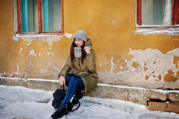 추운 날씨에 오래된 집의 주황색 벽에 회색 스카프와 모자 안경을 쓴 갈색 머리 소녀의 초상화