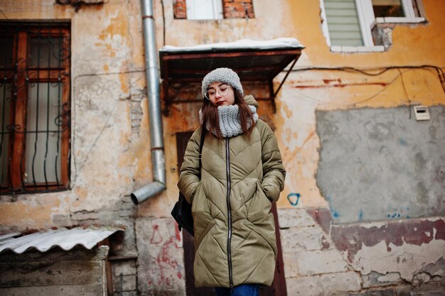 Portrait of brunette girl in gray scarf and hat glasses at cold weather against orange wall of old house