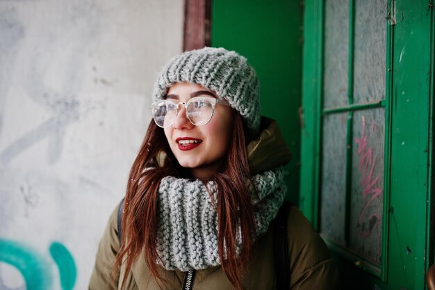 Portrait of brunette girl in gray scarf and hat glasses sitting against urban entrance