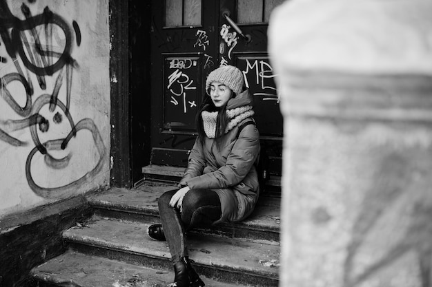 Portrait of brunette girl in gray scarf and hat glasses sitting against urban entrance