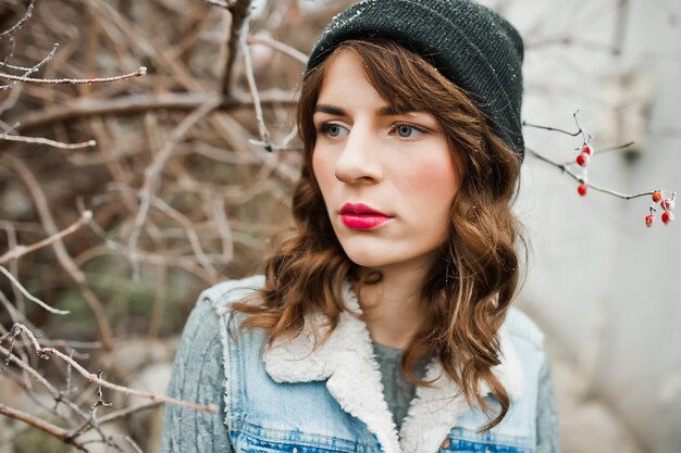 얼어붙은 덤불에서 모자와 청바지 재킷을 입은 갈색 머리 소녀의 초상화