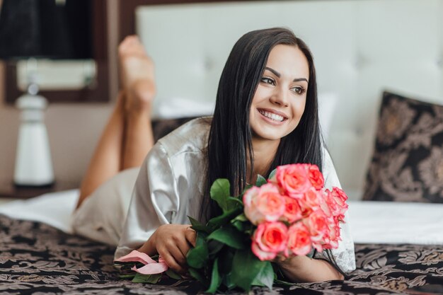 Portrait of brunette girl in the morning in her room, which is lying on a bed with a bouquet of roses in her arms.