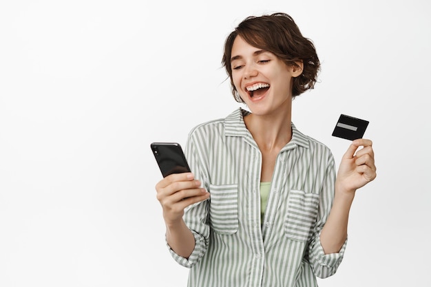 Portrait of brunette girl paying, shopping online with mobile phone and credit card, purchase in app, standing over white background. Copy space