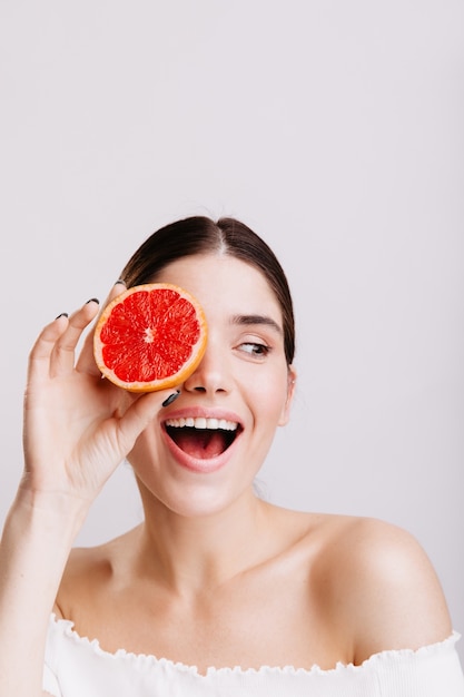 Portrait of brunette girl smiling on white wall. Woman without makeup posing with grapefruit.