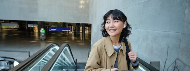 Free photo portrait of brunette girl in wireless headphones listens music uses smartphone goes on escalator