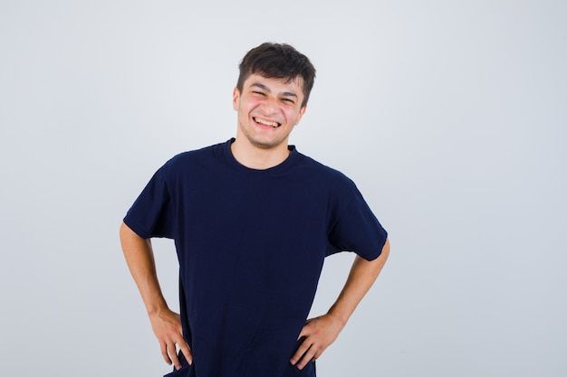 Portrait of brunette man looking at camera, holds hands on waist in dark t-shirt and looking cheerful, front view.