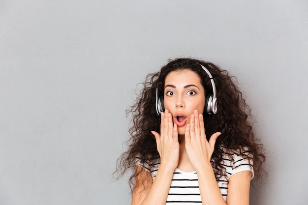 Portrait of brunette mixed race woman listening to music via wireless headphones putting hands to mouth meaning surprise over grey wall
