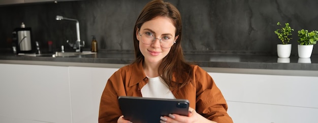 Portrait of brunette woman in glasses sitting in kitchen at home holding digital tablet reading on