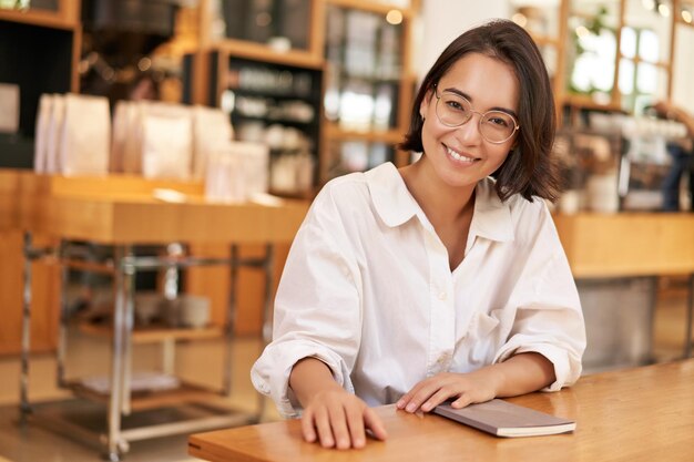 Портрет брюнетки в очках, улыбающейся с уверенным взглядом, сидящей в кафе с ноутбуком