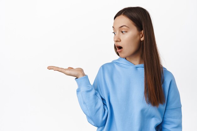 Portrait of brunette woman looking at her palm, empty open hand, holding smth, display at item, advertisement of product on sale, standing against white wall