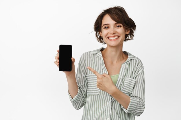 Portrait of brunette woman showing smartphone screen, pointing finger at mobile phone and smiling happy, demonstrating app, standing over white background.