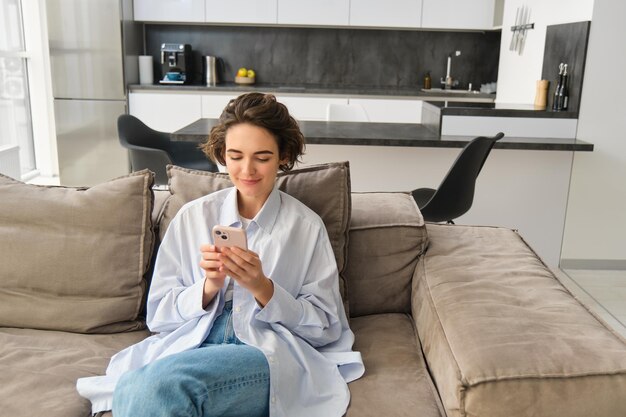 Portrait of brunette woman sits on sofa with smartphone chats on phone looks at her mobile app check