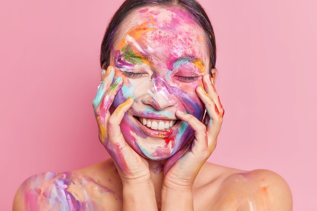 Portrait of brunette young Asian woman smiles pleasantly keeps both hands on cheeks stands with closed eyes has face smeared by colorful oil paints 