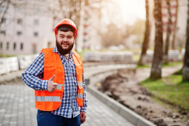 Portrait of brutal beard worker man suit construction worker in safety orange helmet against pavement shows thumb