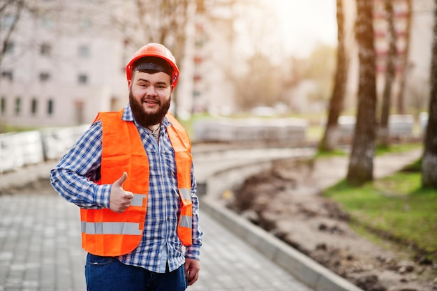 Portrait of brutal beard worker man suit construction worker in safety orange helmet against pavement shows thumb