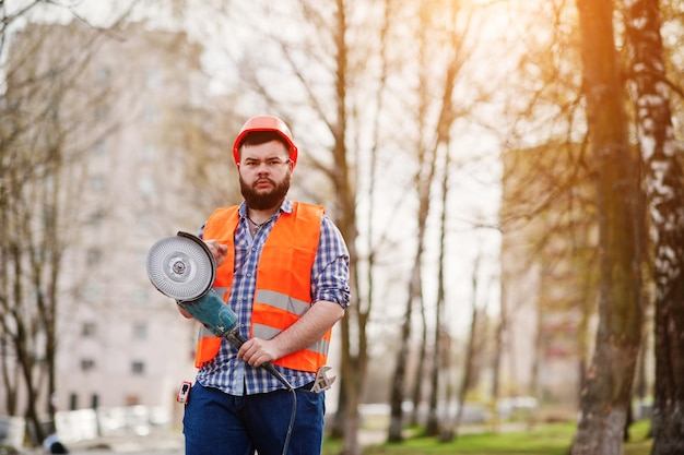 Portrait of brutal beard worker man suit construction worker in safety orange helmet against pavement with angular grinding machine in hand