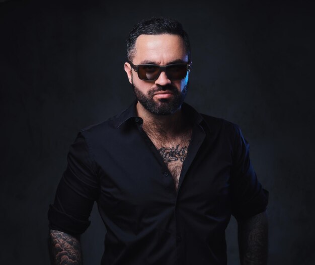 Portrait of brutal bearded male in sunglasses with tattoos on arms and neck.