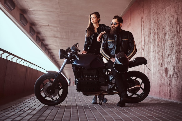 Portrait of a brutal fashionable biker dressed in a black leather jacket with sunglasses sitting on his custom-made retro motorcycle and his young brunette girlfriend who leaning on his shoulder.