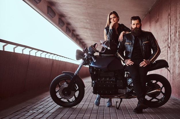 彼のカスタムメイドのレトロなオートバイと彼の肩に寄りかかっている彼の若いブルネットのガールフレンドに座っているサングラスと黒い革のジャケットに身を包んだ残忍なファッショナブルなバイカーの肖像画。