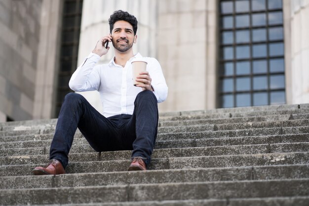 Портрет делового человека разговаривает по телефону, сидя на лестнице на открытом воздухе